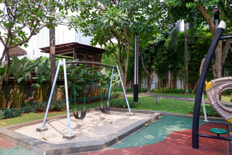 Swing set and timber play house on rubber matting, shaded by tall planting