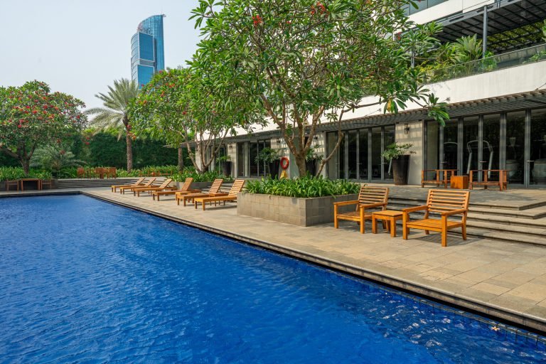 Poolside deck with timber loungers and side tables lining the water's edge, greenery along the far side