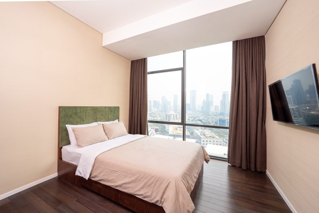 Bedroom with a green upholstered headboard, wall-mounted TV and floor-to-ceiling window onto the Jakarta skyline